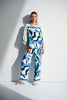 A person models against a white wall in the Joseph Ribkoff Woven Abstract Print Boxy Top 252085, showcasing its blue, green, and white wavy patterns with a chic boat neck. They accessorize with large earrings and high heels as light casts a shadow to the left.