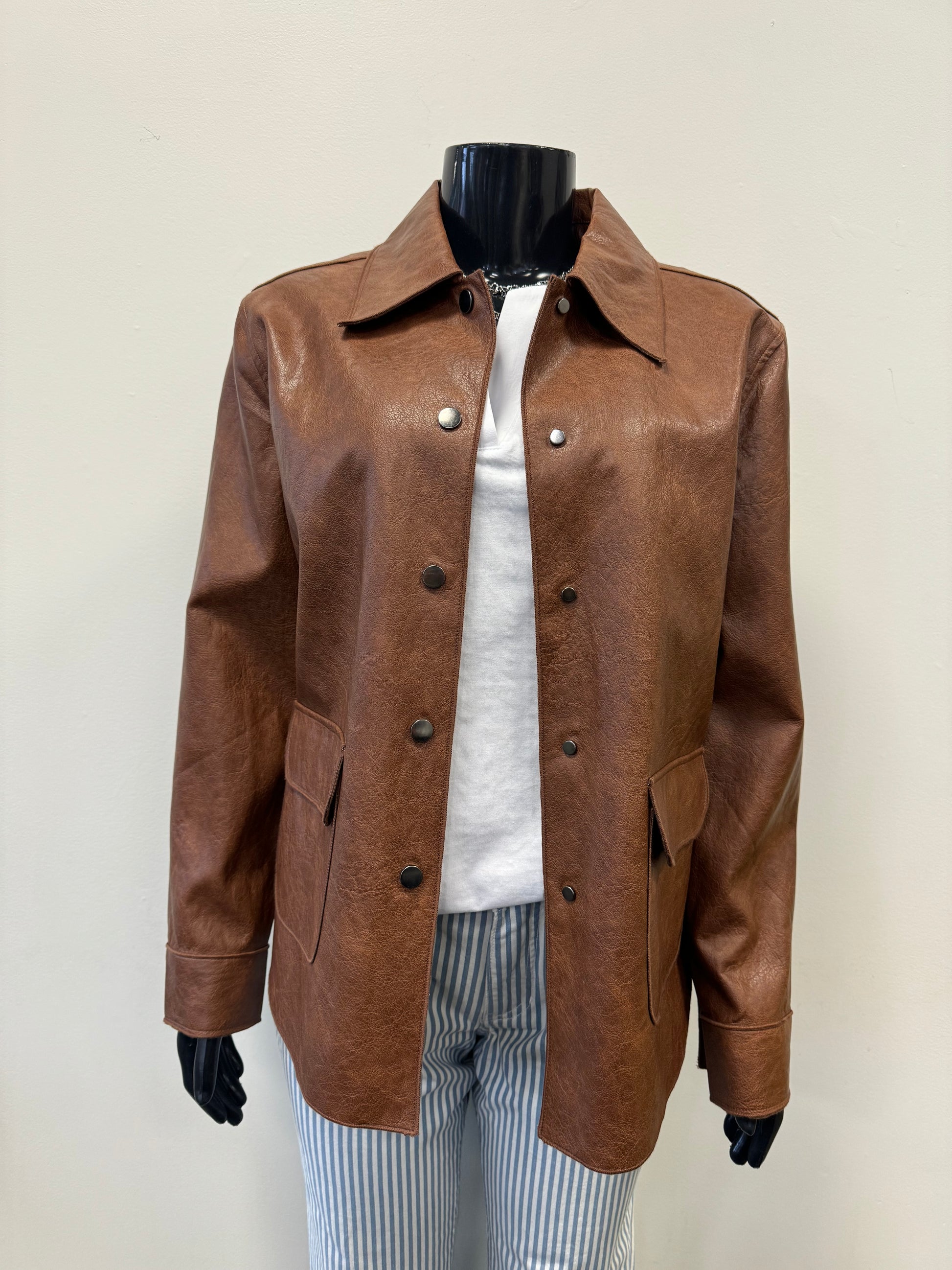 A mannequin wears a stylish and durable Brown Mock Leather Jacket by Tribal over a white T-shirt. The jacket features a collar, buttons down the front, and two large pockets. The mannequin also sports blue and white striped pants. The background is plain and off-white, highlighting the modern design of the outfit.