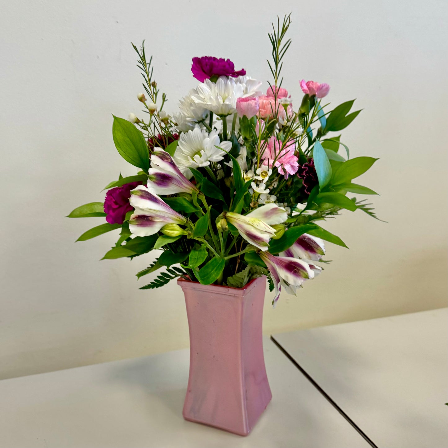 Introducing the "Based in Pink" bouquet by Posies Flowers & Fashion: A bouquet of vibrant flowers arranged in a pink vase. The bouquet includes white daisies, pink carnations, and dark pink carnations, along with assorted greenery. While substitutions may occur based on availability, the same delicate charm is always maintained. The vase sits on a light-colored surface against a plain white background.