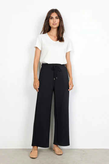 A woman is standing against a plain white background. She has medium-length, straight hair and is wearing a white short-sleeved shirt, BANU 116 Pants in black from SoyaConcept with an elastic waist and wide-leg drawstring design, and beige flats. Her arms are relaxed by her sides.