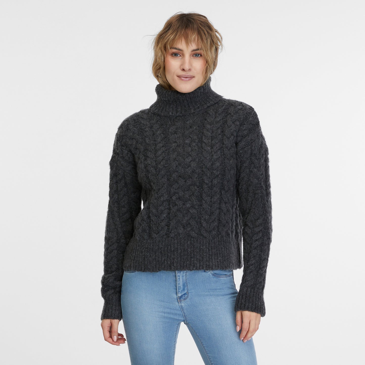 A person with shoulder-length blonde hair stands against a plain white background, wearing the cream-colored Janna Short Flat Knit Sweater by RAGWEAR and light blue jeans. Their relaxed posture and hands loosely hanging by their sides epitomize the comfortable and ethical style of vegan streetwear.