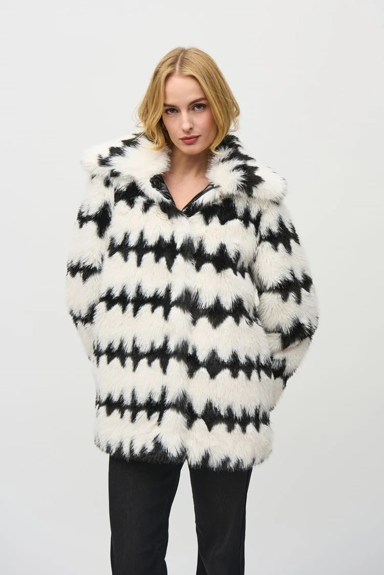 A woman with shoulder-length blonde hair is wearing the Reversible Faux Fur Coat 244900 by Joseph Ribkoff. She has her hands in the coat pockets and stands against a plain white background. The coat features a bold black and white zigzag geometric print.