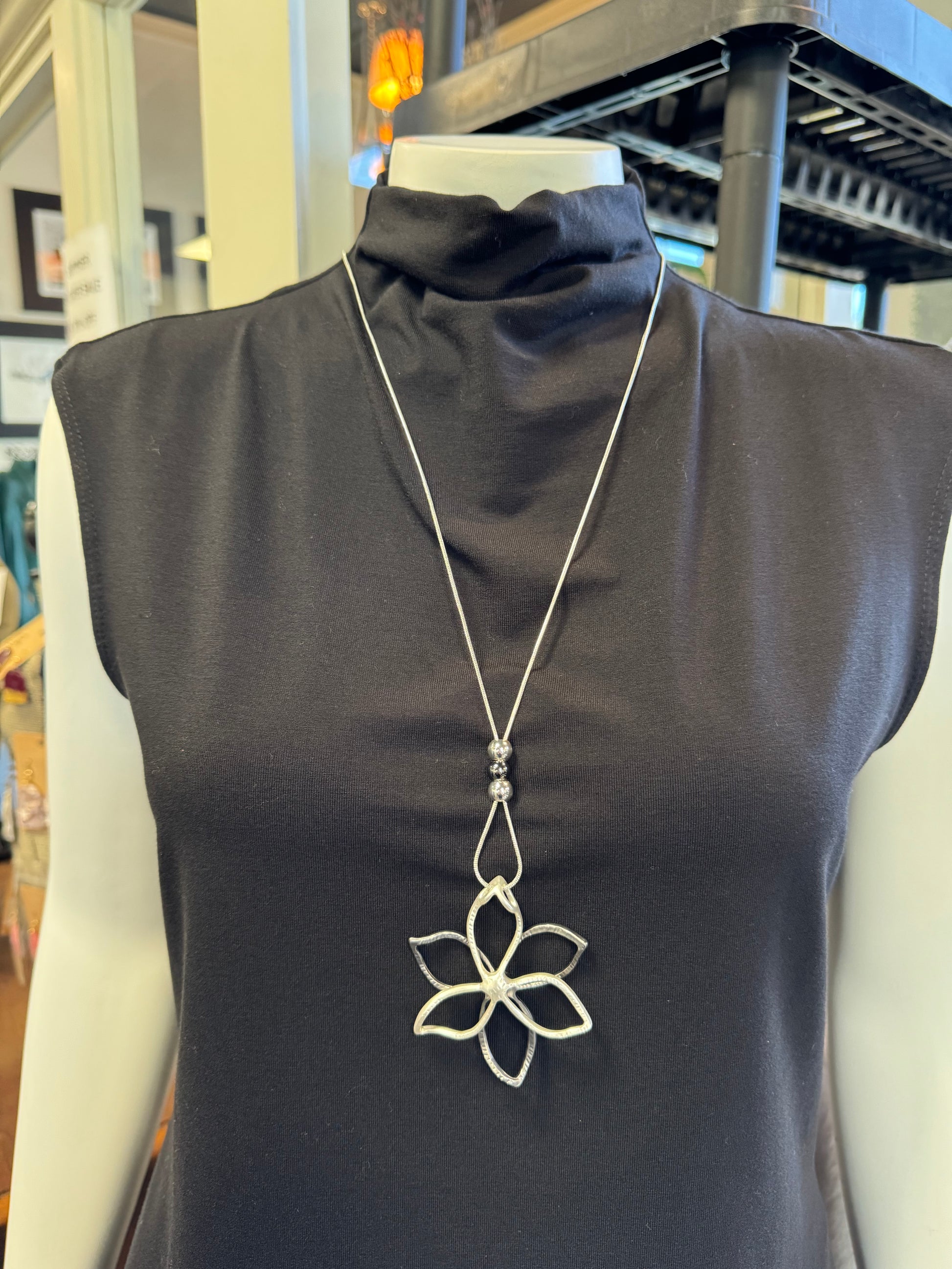 A mannequin in a black sleeveless top is adorned with fashionable jewelry: a long silver necklace from Elise Accessories, showcasing a decorative flower-shaped pendant at its center.