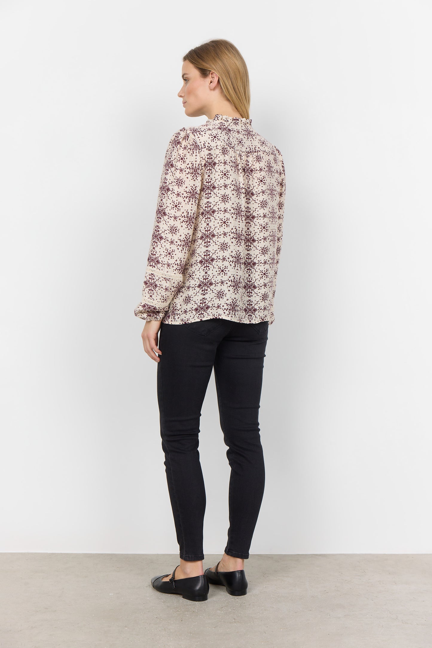 A woman with long blonde hair is wearing the luxurious blend of fabrics in the Shirt SC-IANE 2 from RAGWEAR, featuring a cream-colored blouse with a maroon floral pattern and black pants. She stands against a white background, looking directly at the camera with a neutral expression.