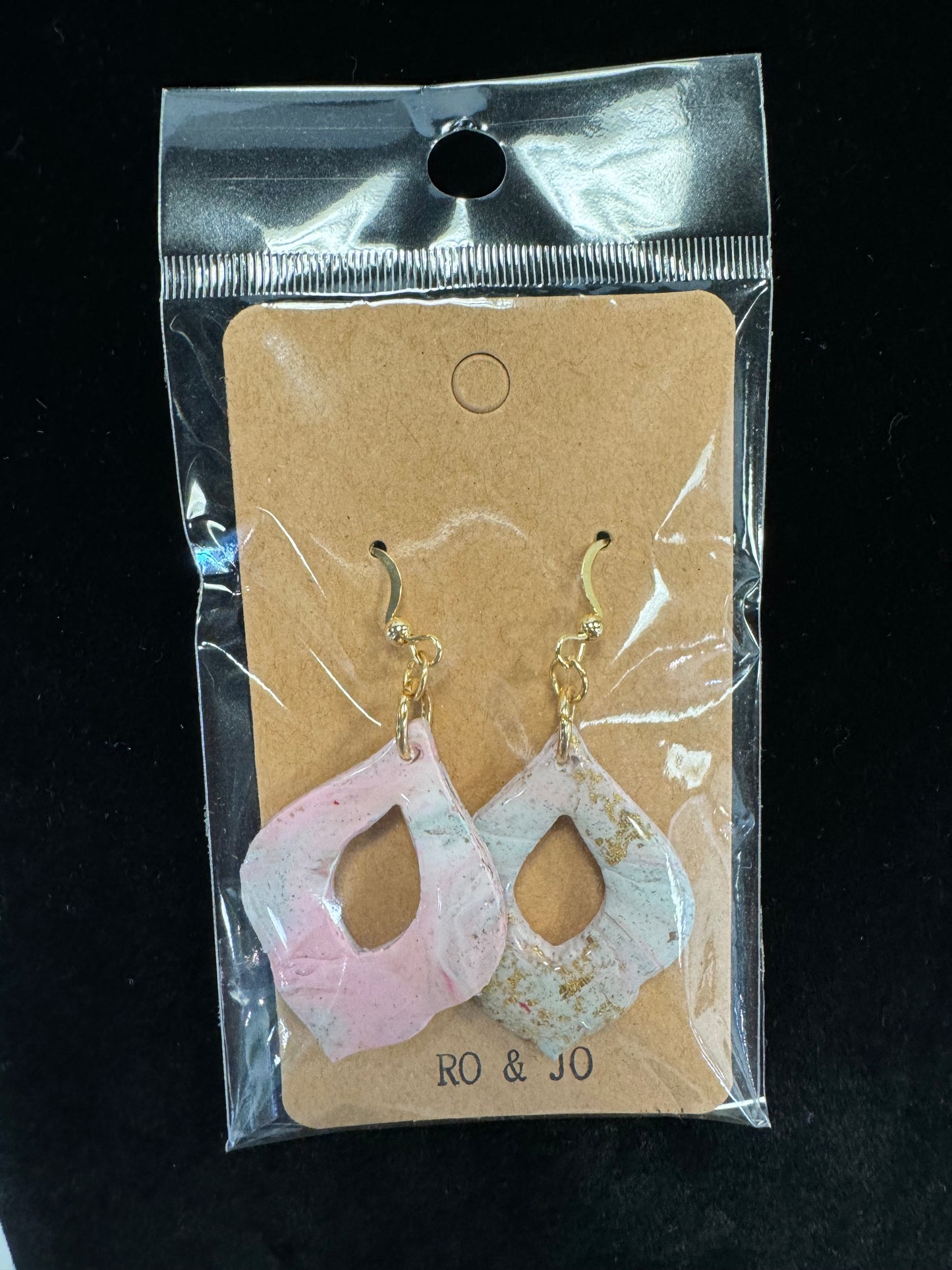 A pair of unique, handmade Ro&Jo Earrings 423 displayed on a brown card with the brand name "RO&JO." The polymer clay earrings feature marbled pink and white designs with gold hooks. The product is packaged in a clear plastic bag against a black background.