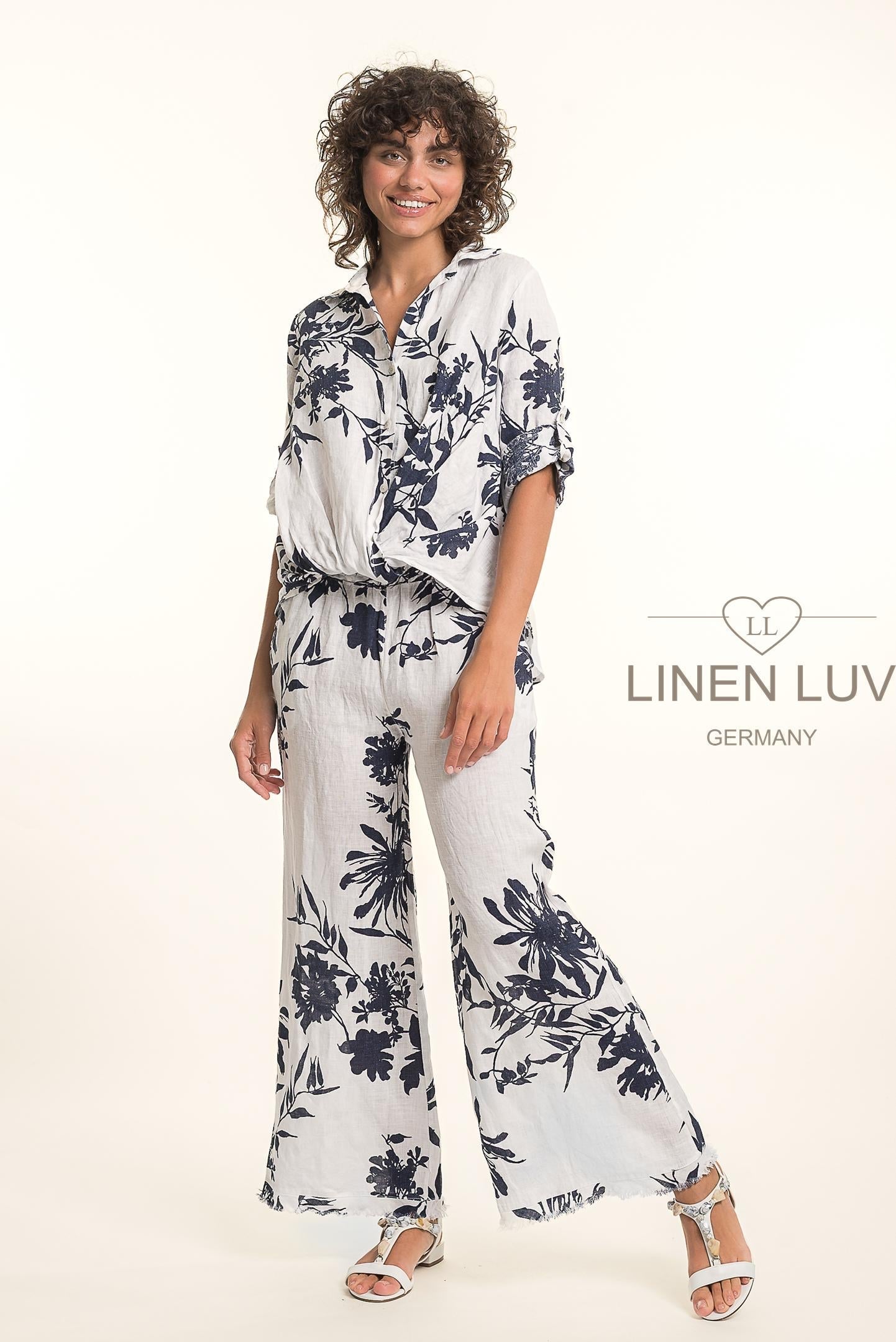 A woman with curly hair wears the Linen Luv French Linen Shirt TP1699P-11 Bianco and matching pants with a dark floral pattern, smiling with one hand in her pocket, sandals on, and the LinenLuv logo to the right.