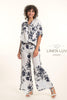 A woman with curly hair wears the Linen Luv French Linen Shirt TP1699P-11 Bianco and matching pants with a dark floral pattern, smiling with one hand in her pocket, sandals on, and the LinenLuv logo to the right.