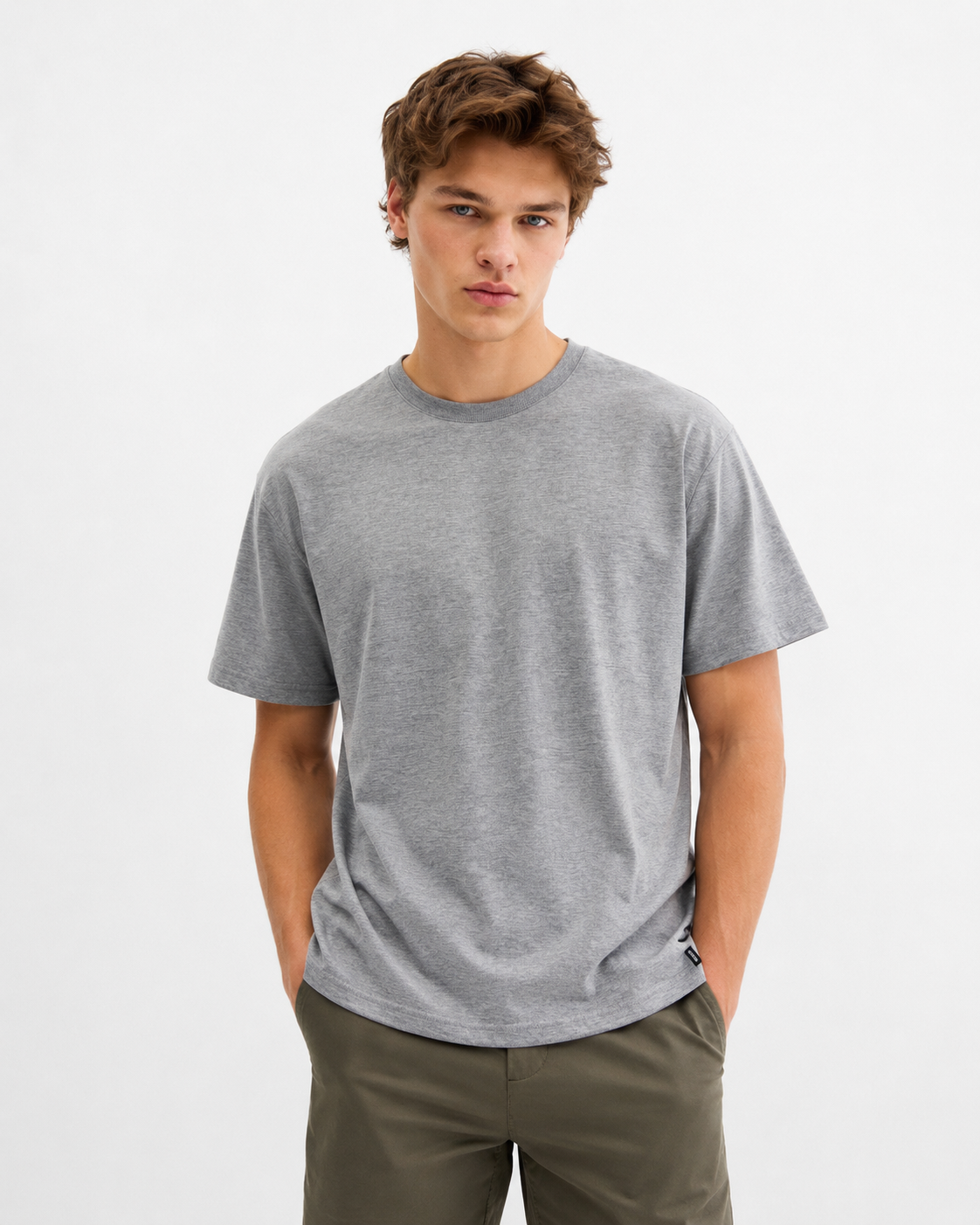 A young man with tousled brown hair wears the RAGWEAR NEDDIS 2512-15002-3000 T-shirt in grey and olive green pants, standing with his hands in his pockets against a light plain background.