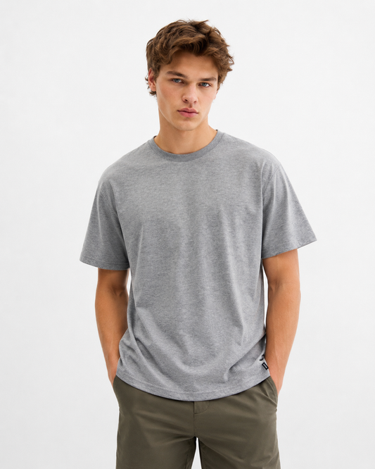 A young man with tousled brown hair wears the RAGWEAR NEDDIS 2512-15002-3000 T-shirt in grey and olive green pants, standing with his hands in his pockets against a light plain background.