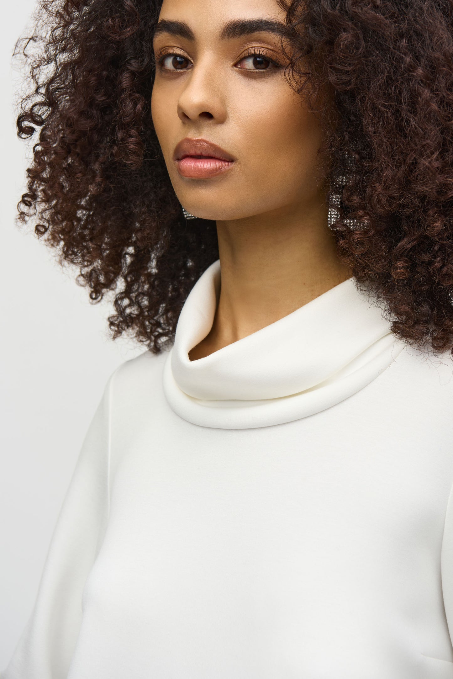 A woman with curly hair wears a loose-fitting, long-sleeved white Scuba Jersey Boxy Top 244129 by Joseph Ribkoff. The top, featuring button details on the sleeves, lends a boxy silhouette. She stands in front of a plain background, looking slightly downward with a neutral expression.