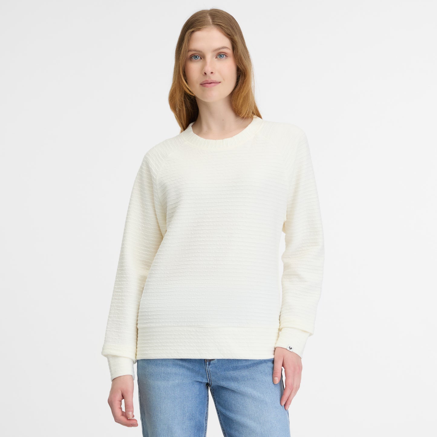 Against a plain white background, a person with long hair wears the JOANN CREPE TOP in cream by RAGWEAR, paired with blue jeans. They look straight ahead with a neutral expression, embodying casual simplicity.