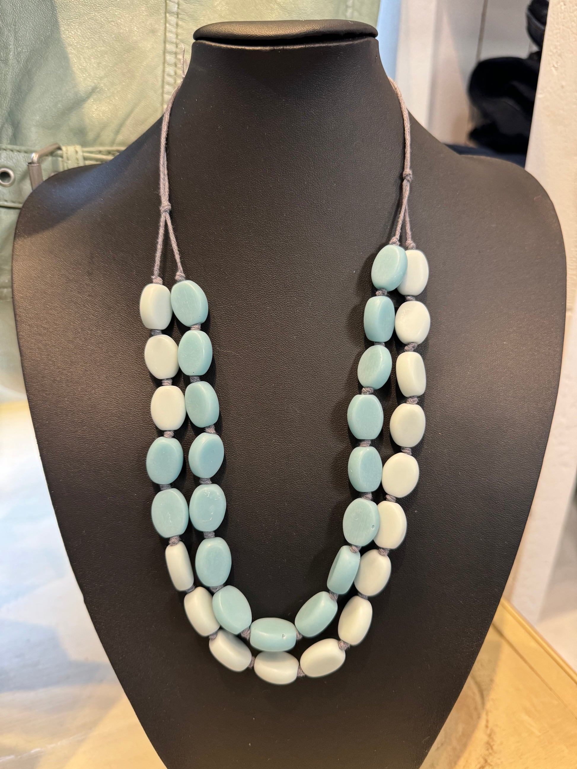 A black mannequin bust displays an elegant accessory: the Suzie Blue Necklace Adjustable in BLUE/GREY. This double-strand necklace, composed of large oval beads alternating in shades of pale blue and white, rests gracefully against the dark background, accentuating its soft pastel and blue-grey hues.