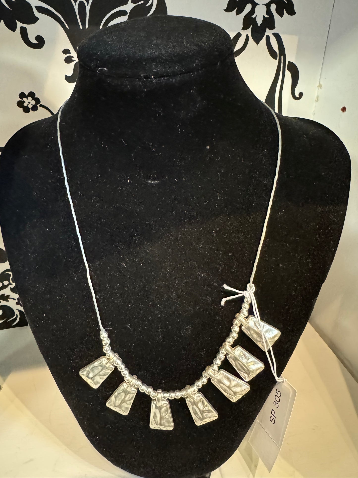 A Suzie Blue black velvet necklace display stand holds an elegant Silver Coloured Necklace with a white cord. This timeless design features a series of rectangular, decorated pendants interspersed with small round beads. A price tag labeled "SP 305" is attached to the necklace.