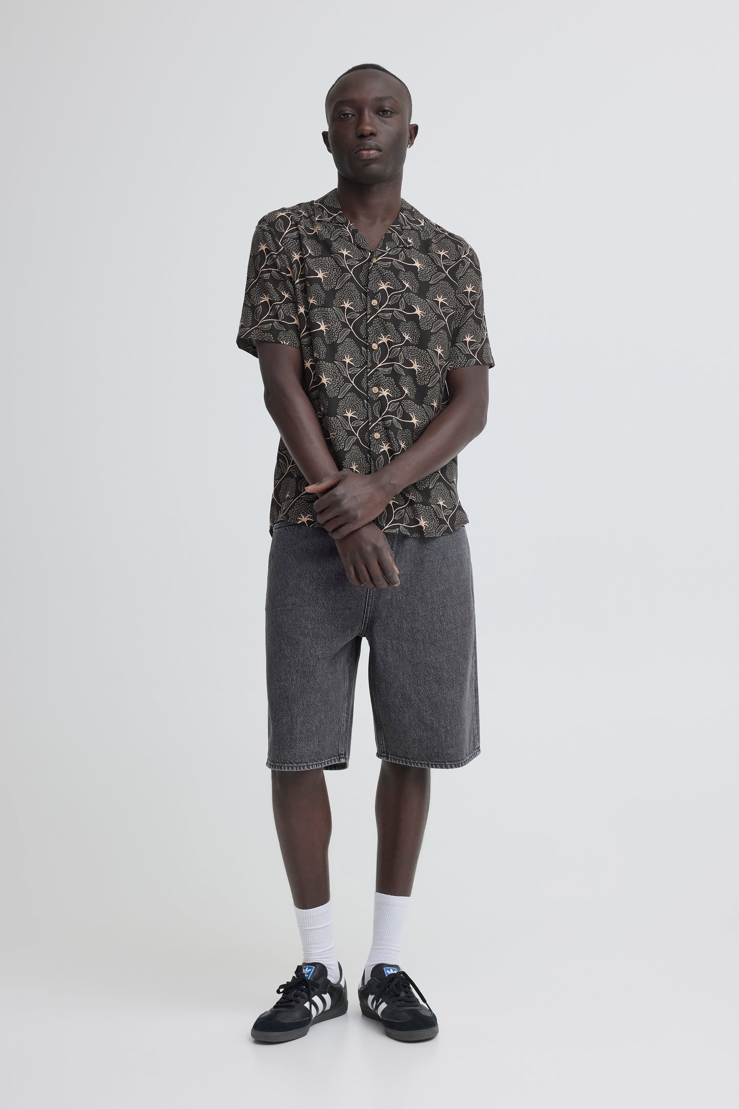 A man stands against a white background wearing the BLEND ARIAN SHIRT 20720641-099996162, gray knee-length shorts, white socks, and black sneakers with white soles. His hands are clasped in front of him.
