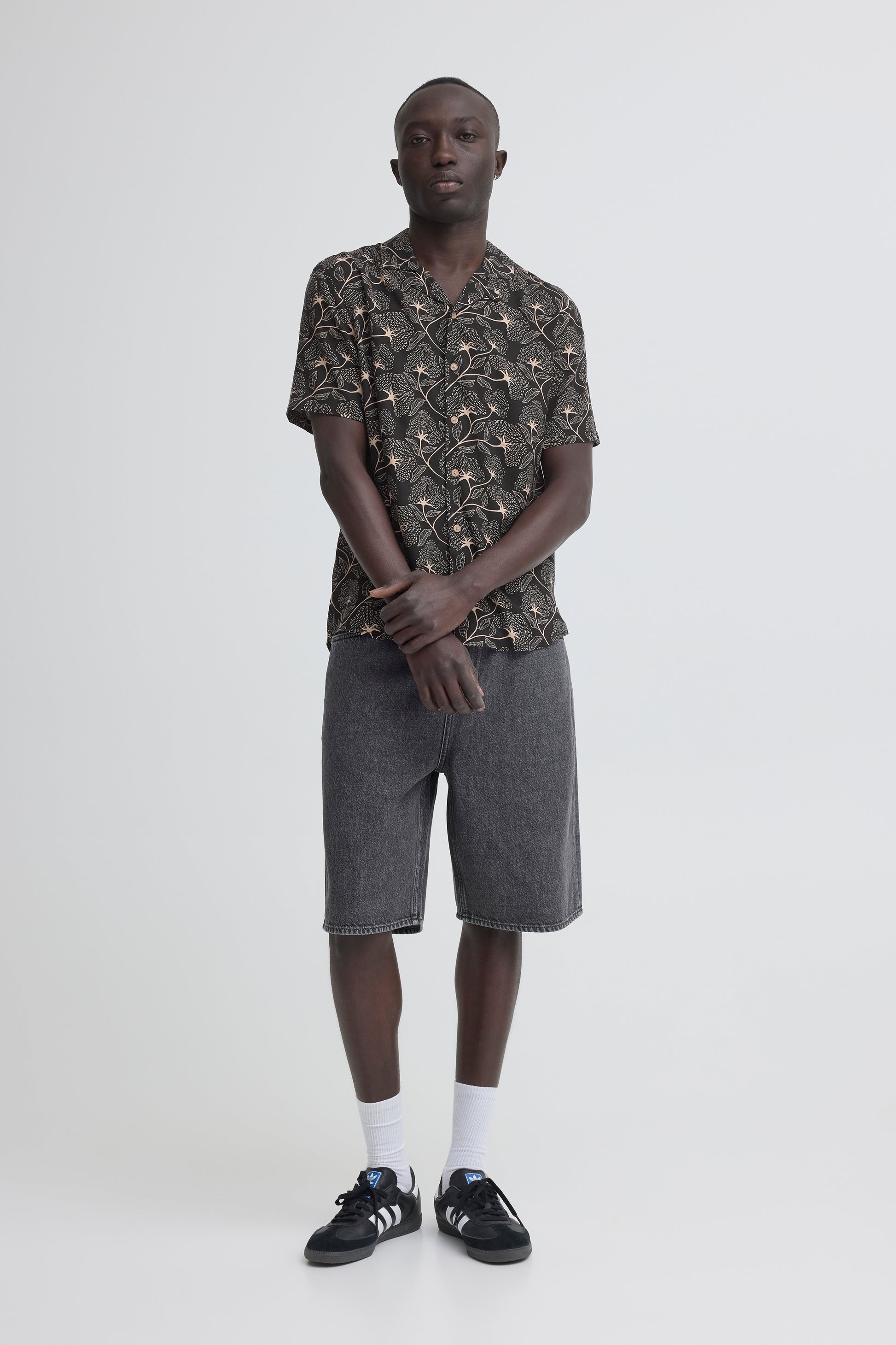 A man stands against a white background wearing the BLEND ARIAN SHIRT 20720641-099996162, gray knee-length shorts, white socks, and black sneakers with white soles. His hands are clasped in front of him.