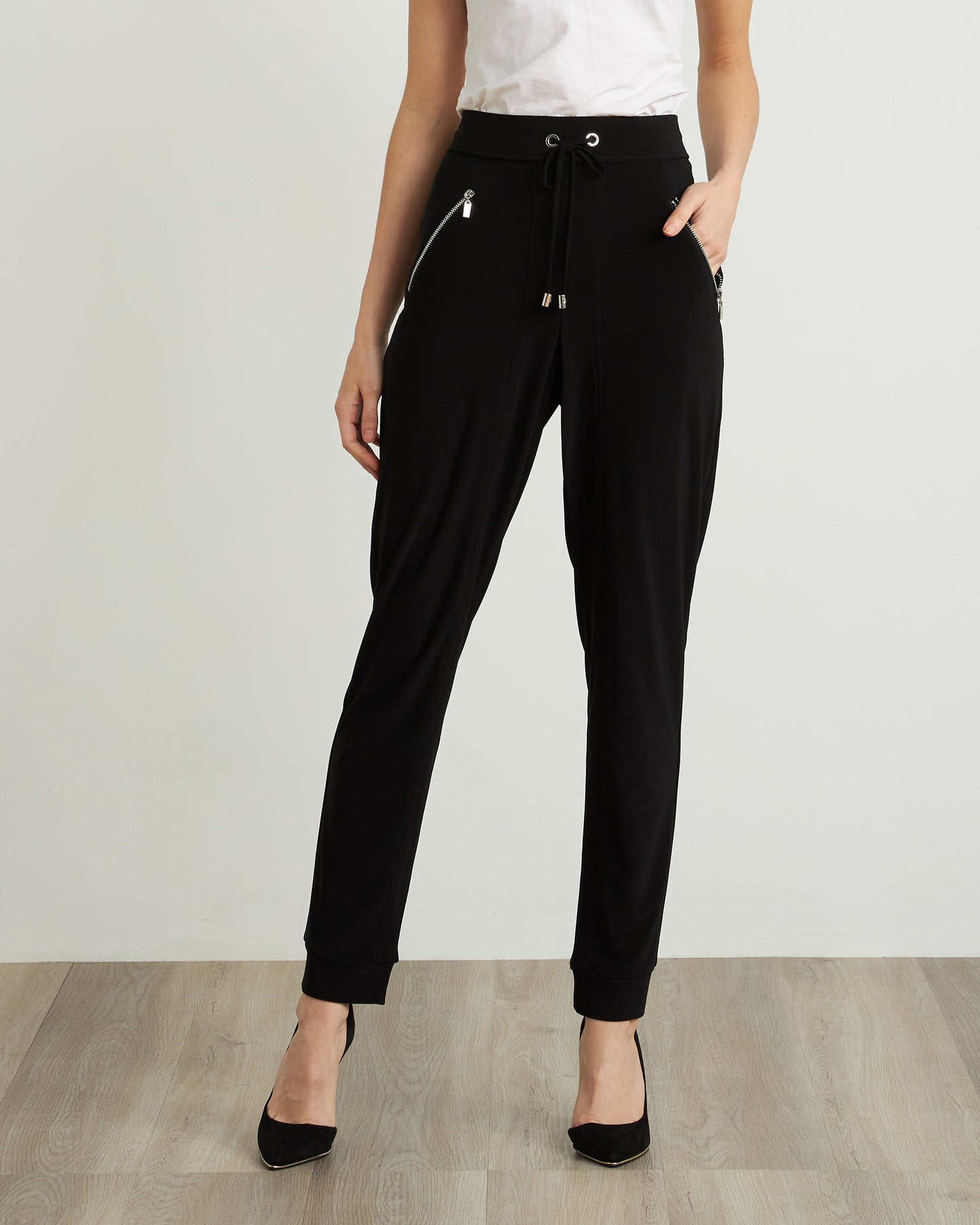 An individual wearing Joseph Ribkoff's Drawstring Waist Pants Style 211317, featuring zipper detailing in black, paired with a white top and black heels. The person stands on a wooden floor against a plain, light-colored background, seamlessly combining comfort and style.