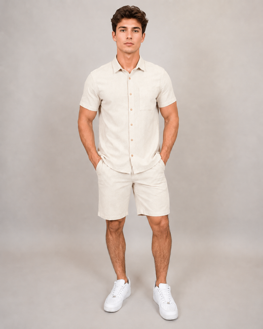 A man stands with hands in pockets, wearing the HEDGE Linen Blend Short Sleeve Button Shirt 56MW104S and matching beige shorts with white sneakers—an ideal warm-weather look—against a plain light grey background.