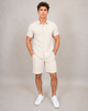 A man stands with hands in pockets, wearing the HEDGE Linen Blend Short Sleeve Button Shirt 56MW104S and matching beige shorts with white sneakers—an ideal warm-weather look—against a plain light grey background.