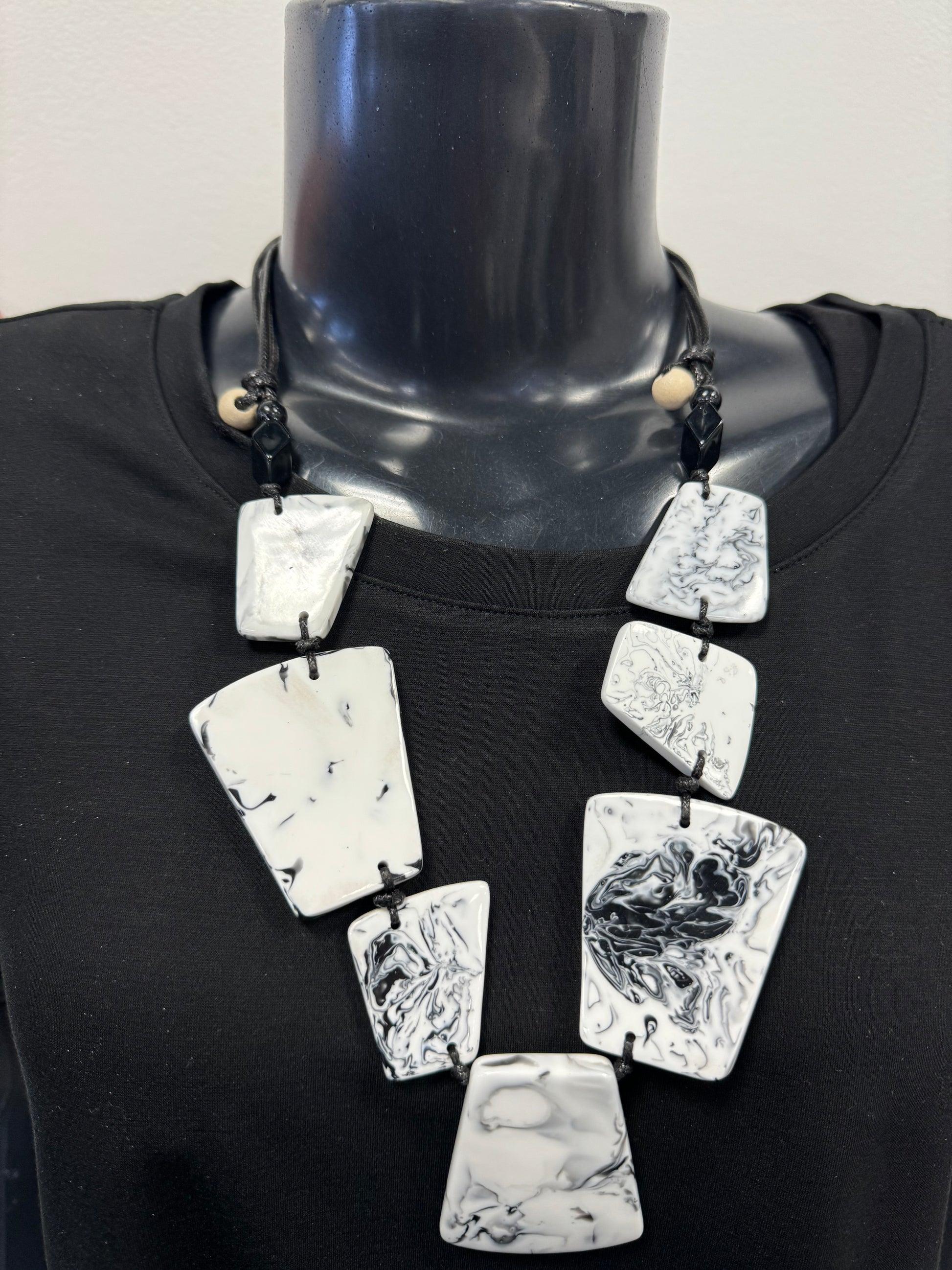 A black mannequin wearing a black garment is adorned with the Posies Flowers & Fashion Inc. White and Black Adjustable necklace, which features large, irregularly shaped white stones with a marbled look interconnected by black cords. Two small wooden beads are visible near the top of the necklace.