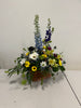 A basket from Posies Flowers & Fashion, named Natural Daisies Basket, showcases a vibrant arrangement of blue delphiniums, white and yellow daisies, and green ferns. The flowers are artfully displayed on a white surface against a plain wall background.