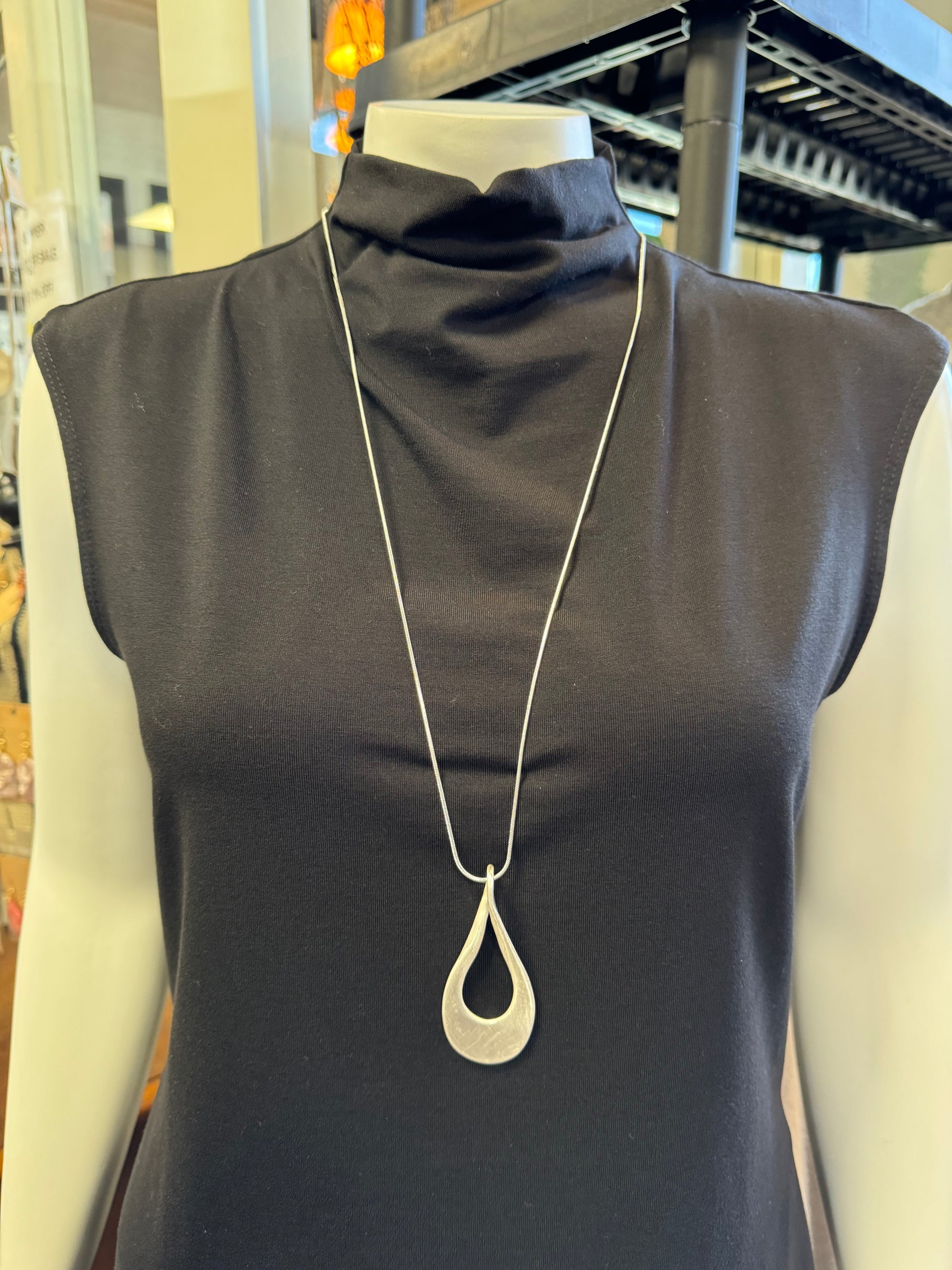 A mannequin wearing a black sleeveless turtleneck top is highlighted by the stunning **Andy 19 Necklace** from this season's jewelry collection by Elise Accessories, featuring a long silver chain and teardrop-shaped pendant. The background includes some indoor decor elements, enhancing this exquisite fashion piece.