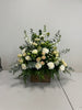 The White Sympathies Arrangement from Posies Flowers & Fashion features a wooden container that holds a lush assortment of white and cream flowers, including roses, carnations, and chrysanthemums. Green foliage and eucalyptus leaves extend gracefully from the sides, set against a plain white background. Please check availability for this elegant floral display.