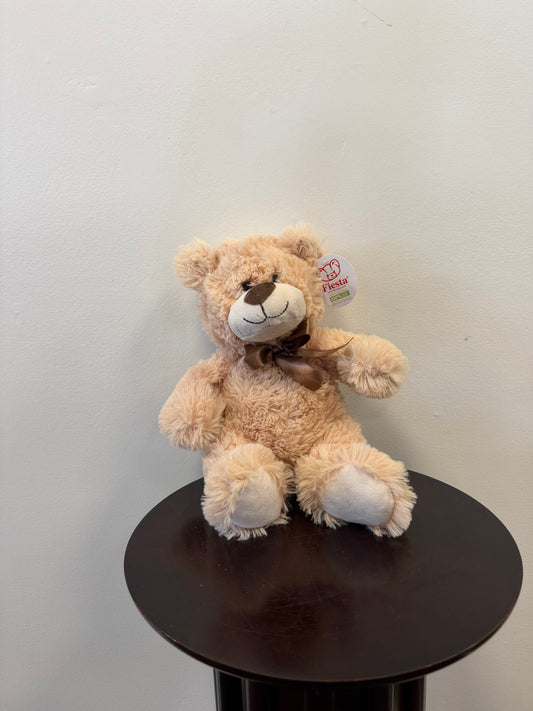A Classic Teddy Bear 9 inch – Fiesta by Fiesta with a brown ribbon sits on a dark round table against a plain white wall. This plush teddy bear features a tag on its ear and a smiling face, making it an ideal gift delivery choice for any occasion.