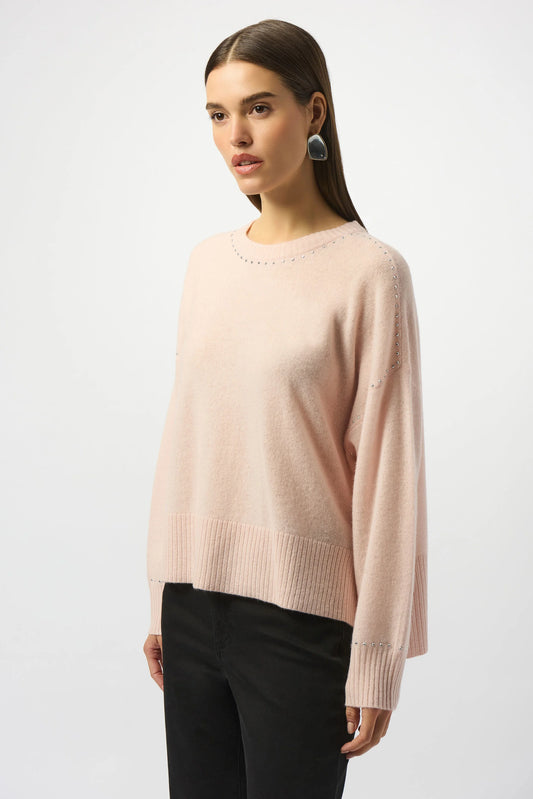 A woman with straight brown hair wears the Joseph Ribkoff Cashmere Crew Neck Top with Stud Detailing 253980 and dark pants, standing against a plain light background and looking slightly to the side.