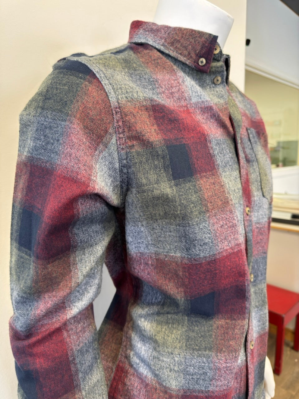 The mannequin is showcasing the Shirt Long Sleeve - BURGUNDY, a button-up flannel with a plaid pattern in shades of red, green, gray, and blue. Expertly paired with denim from Silver Jeans Co., this shirt boasts a single chest pocket and is displayed with the sleeves down and buttons fastened.