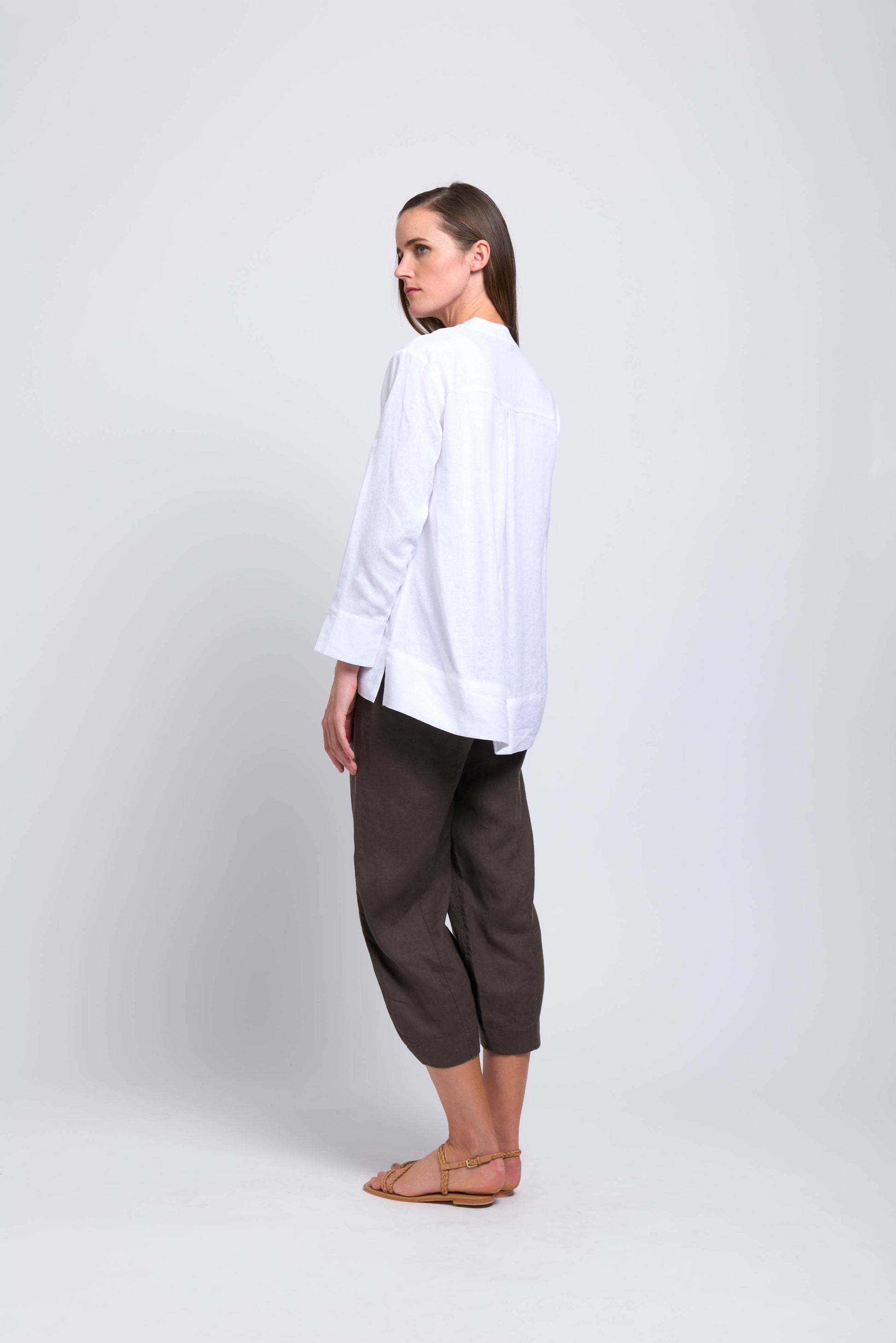 A woman faces away, glancing over her shoulder. She wears the FOIL Artful Ease Shirt FO7906 White with dark cropped pants and sandals, set against a plain light background.