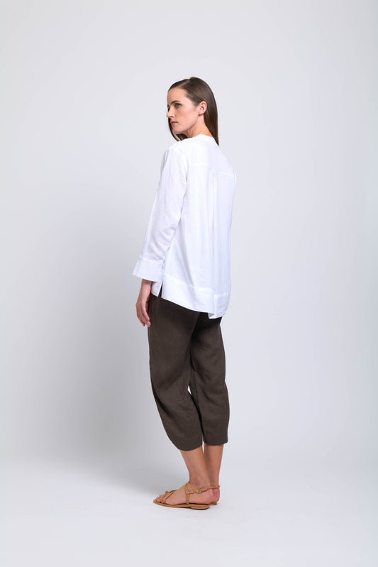 A woman faces away, glancing over her shoulder. She wears the FOIL Artful Ease Shirt FO7906 White with dark cropped pants and sandals, set against a plain light background.