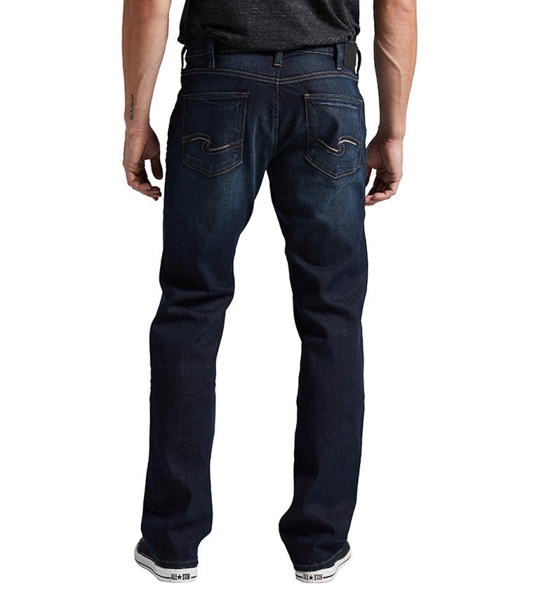 A person wearing Allan Slim Fit Straight Leg Jeans by Silver Jeans Co., a dark shirt, and black-and-white sneakers stands against a plain white background. The person's arms hang naturally by their sides, with the left arm slightly bent. Only the lower half of the body is visible.