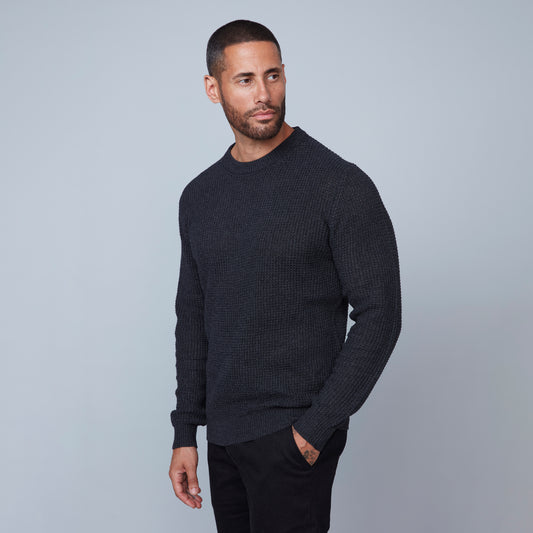 A man with short hair and a beard stands against a plain gray background. He is wearing a dark gray, long-sleeved Crewneck Sweater from HEDGE, made from recycled cotton and black pants, looking slightly to his left. His right arm hangs by his side, and his left arm is bent at the elbow.