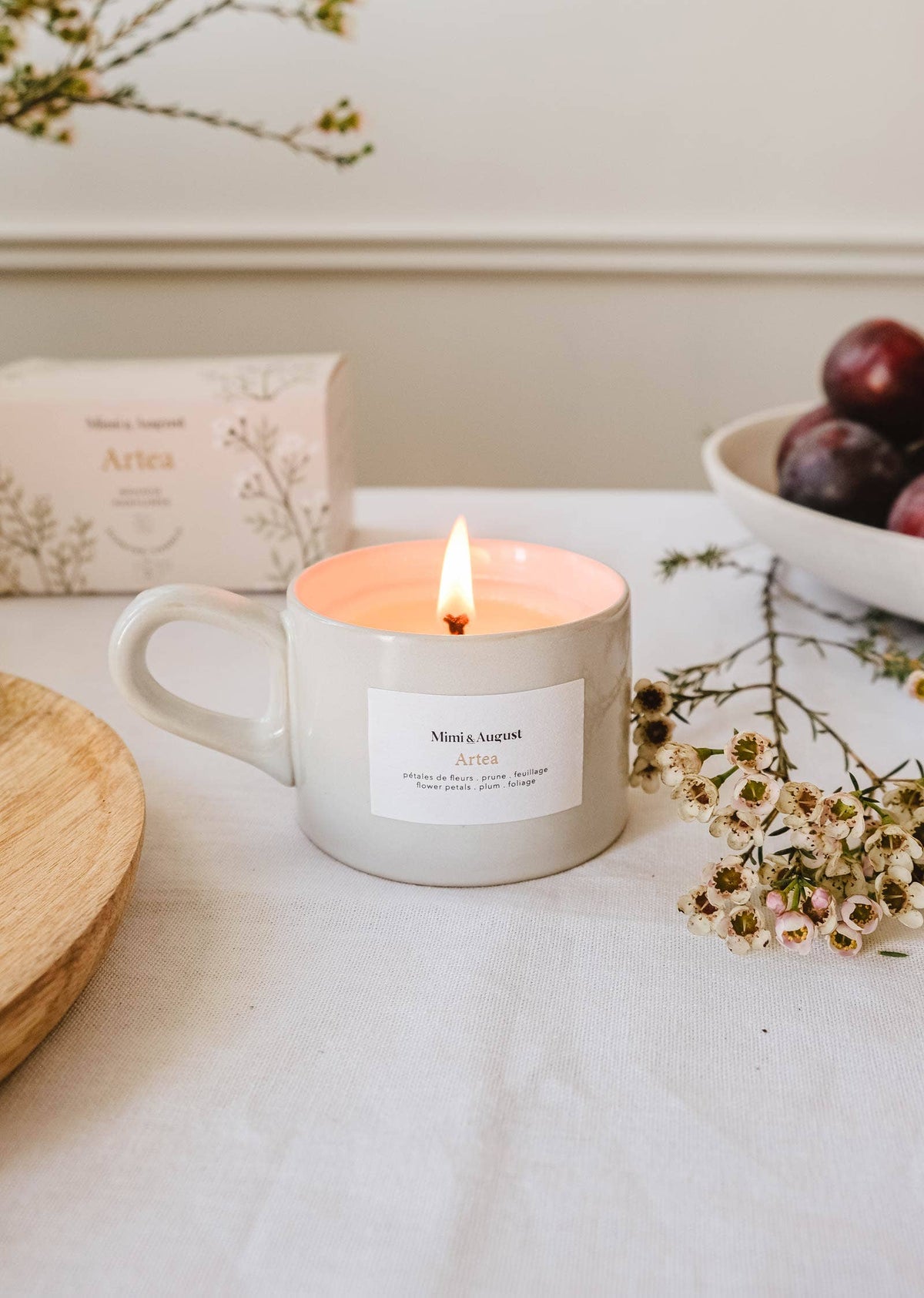 A lit Mimi & August Artea reusable candle (5.5 oz) in a white mug sits on a table next to a bowl of plums, a box, a wooden plate, and sprigs of small white flowers.