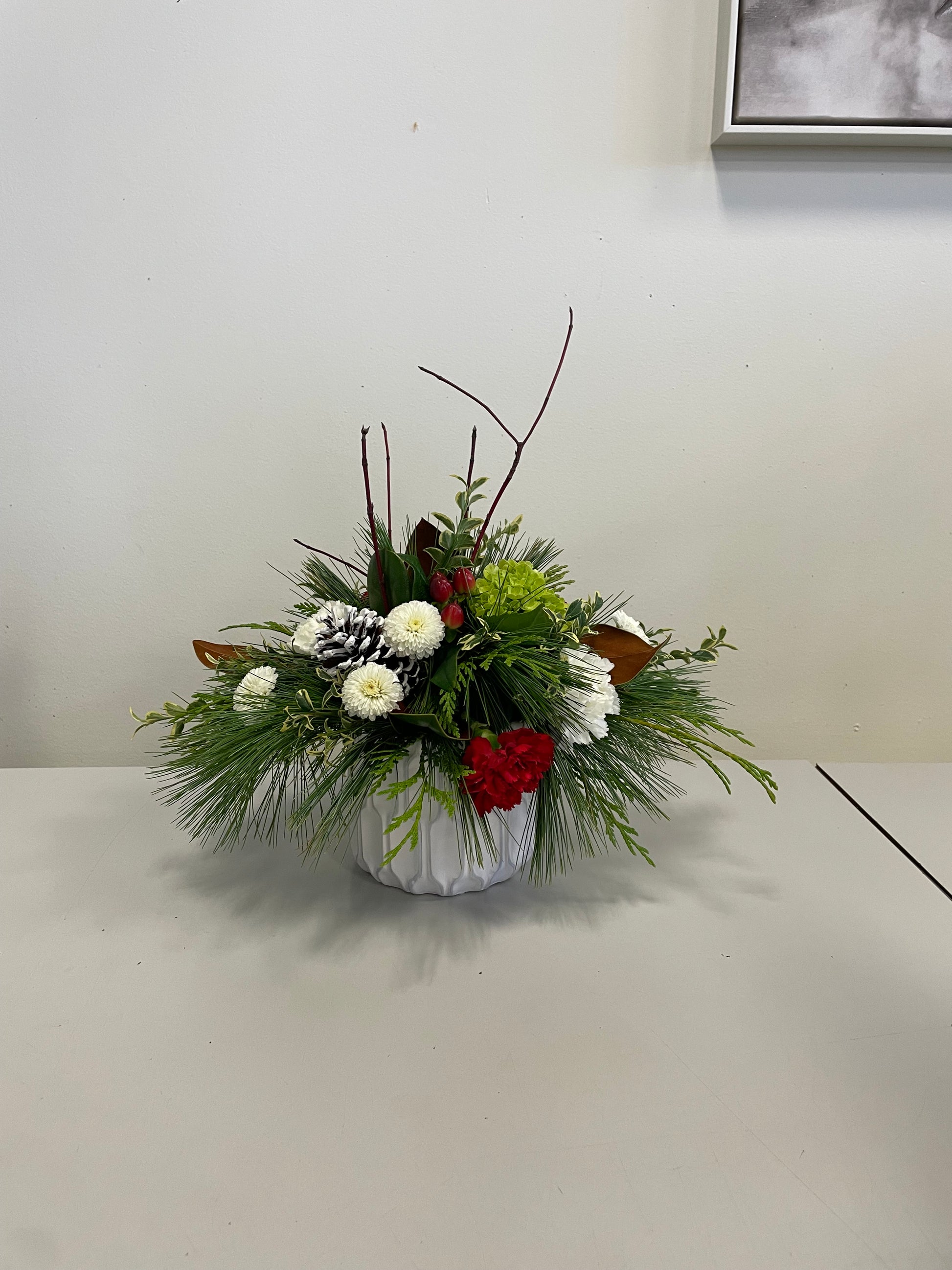 The Winter Fresh Centrepiece from Posies Flowers & Fashion is beautifully displayed on a table, showcasing white and red flowers, green foliage, and two pine cones in a white ceramic vase, exuding festive charm that makes for the perfect holiday decor.