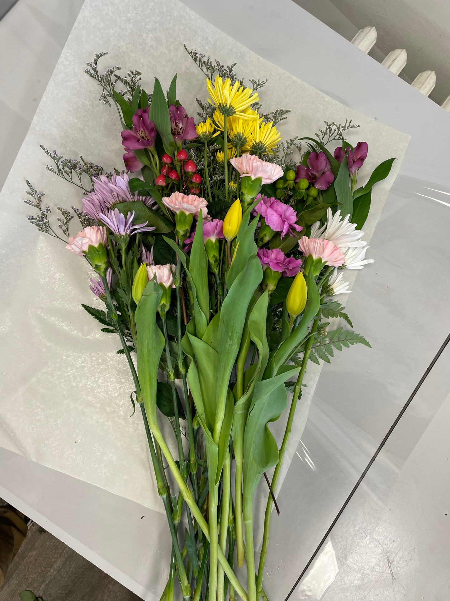 A stunning bouquet called Designer’s Choice Freshcuts from Posies Flowers & Fashion is resting on a white surface. The arrangement features vibrant orange and pink roses, cheerful yellow chrysanthemums, elegant white flowers, and assorted green foliage. It is partially wrapped in brown paper and elegantly tied with a burgundy ribbon.