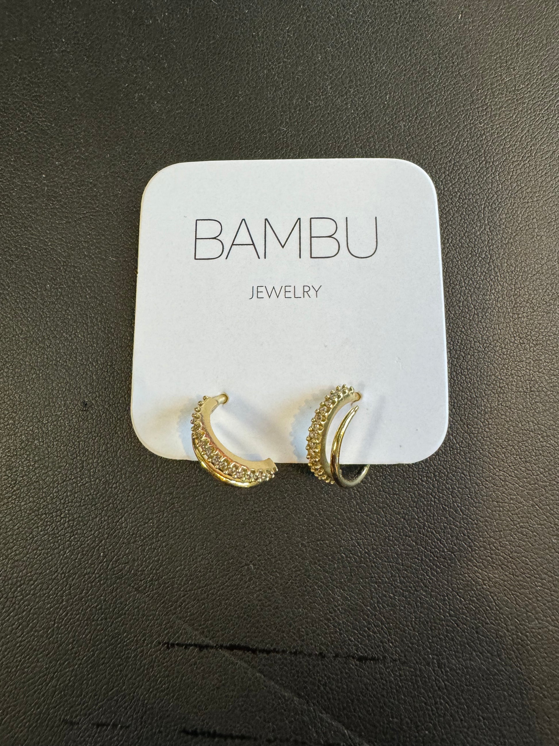 A stunning pair of Gold Embezzled Hoop Earrings from BAMBU Jewellery adorns a white card with "BAMBU JEWELLERY" in gray. These elegant earrings are gold-colored and feature small, clear stones along the front. The card and earrings rest against a dark, textured surface.