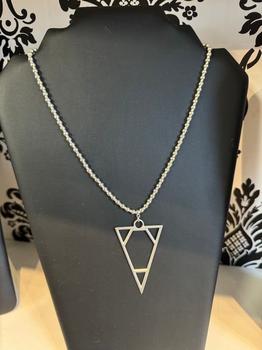 The Suzie Blue Silver Coloured Triangle Necklace, featuring a geometric pendant with an elongated triangle design, is elegantly displayed on a black mannequin. The pendant hangs from a chain made of small silver beads, exuding timeless jewelry charm against the backdrop of a black and white ornate pattern.