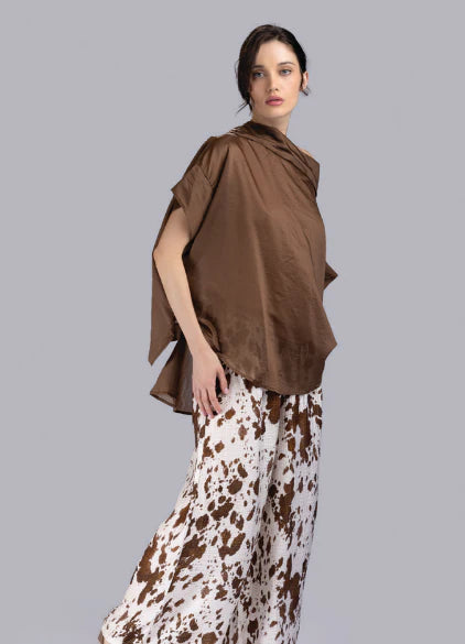 A woman wears the Top FD018 Coco by Paolo Tricot, a loose Tencel top with a draped neckline, paired with wide-legged white pants featuring a brown abstract splatter pattern, standing against a plain gray background.