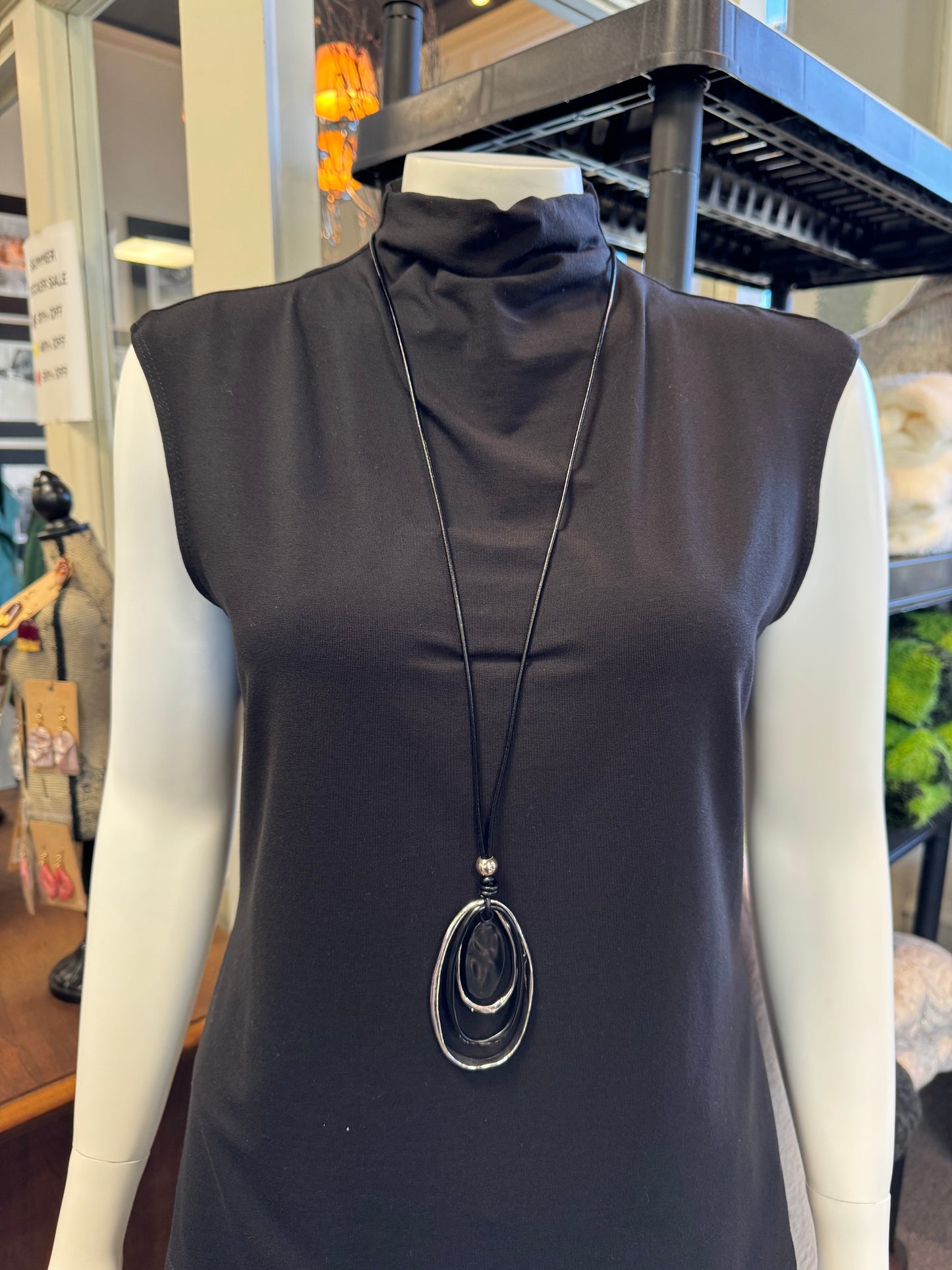 A mannequin displays a sleeveless black turtleneck top in-store, elegantly accessorized with Necklace 4 by Elise Accessories. This long black necklace features a pendant of three oval shapes, making it the perfect fashion accessory. In the background, shelves and assorted items complete the stylish display.