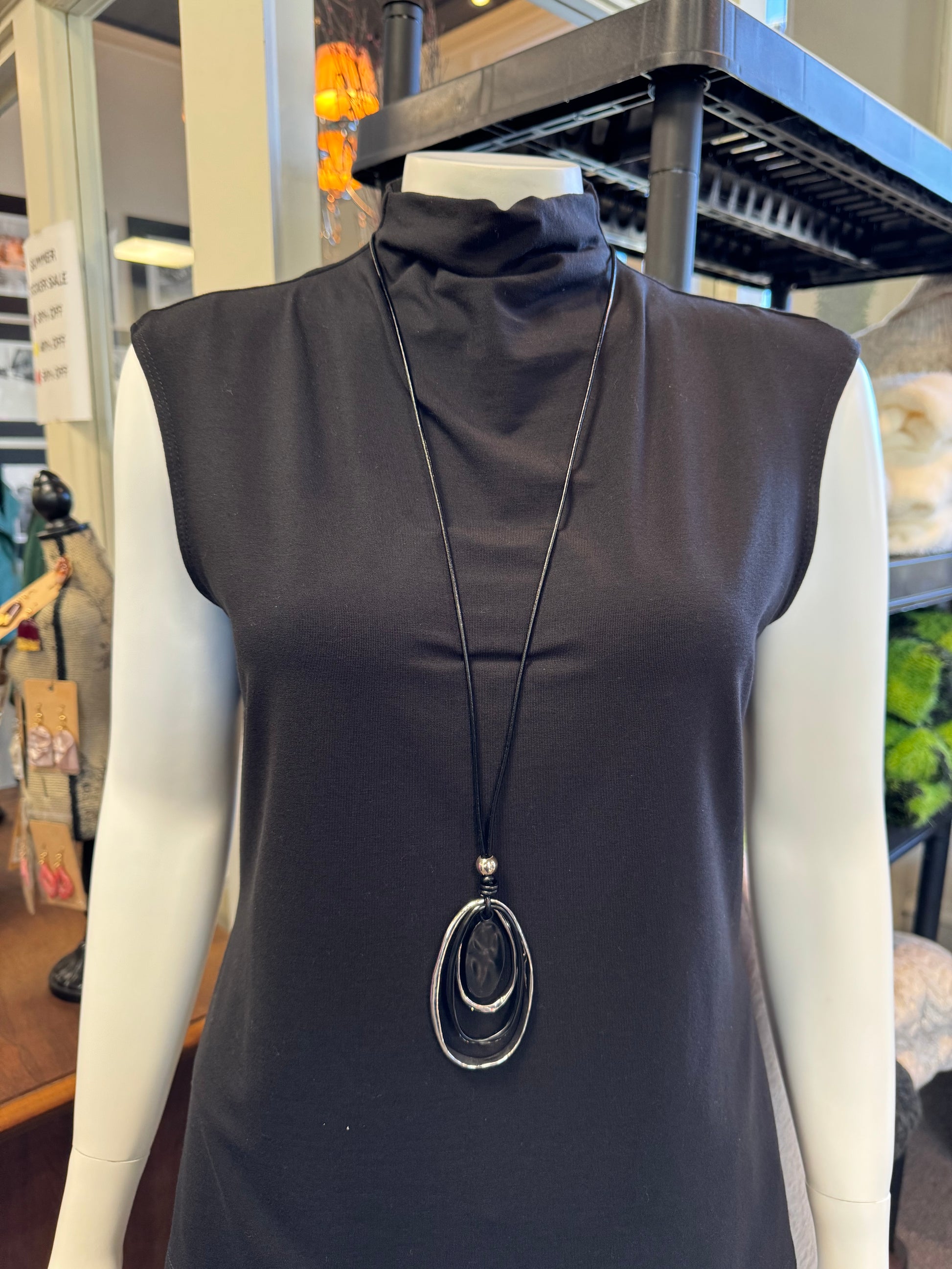 A mannequin displays a sleeveless black turtleneck top in-store, elegantly accessorized with Necklace 4 by Elise Accessories. This long black necklace features a pendant of three oval shapes, making it the perfect fashion accessory. In the background, shelves and assorted items complete the stylish display.