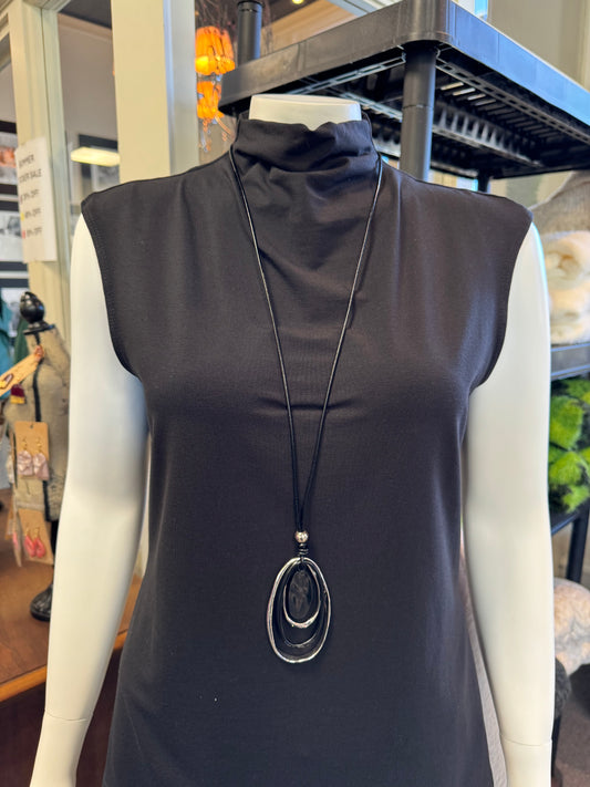 A mannequin displays a sleeveless black turtleneck top in-store, elegantly accessorized with Necklace 4 by Elise Accessories. This long black necklace features a pendant of three oval shapes, making it the perfect fashion accessory. In the background, shelves and assorted items complete the stylish display.