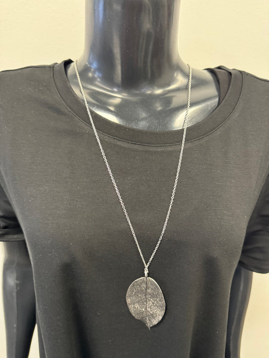 A black mannequin torso wears a black short-sleeve t-shirt and a long silver-colored chain featuring Posies Flowers & Fashion Inc.'s Dark Grey Coloured Leaf Pendant Costume Necklace. The background is plain and light-colored.