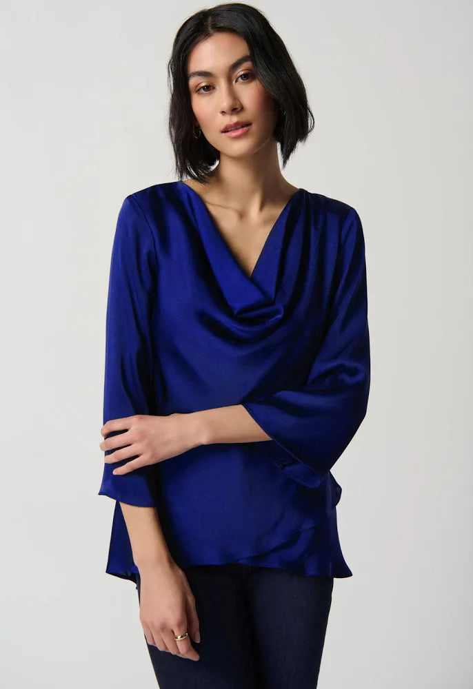 A woman with shoulder-length dark hair wears the Joseph Ribkoff Draped Satin Blouse Style 234082 in Royal Sapphire, featuring long sleeves and a tiered hem, paired with dark pants. She stands against a plain light background with a neutral expression.