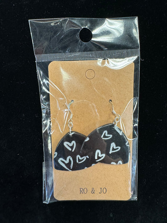 A pair of Ro&Jo Earrings 431, black and heart-shaped with white heart patterns, are displayed in clear plastic packaging. The unique earrings are attached to a brown card with "RO&JO" printed at the bottom. The background is a plain dark surface.