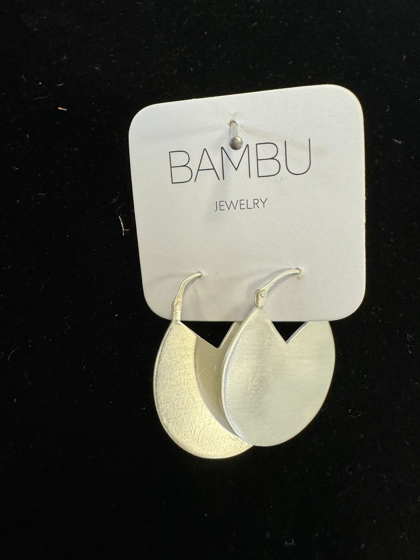 A pair of silver-colored circular earrings hanging on a white rectangular card with a small hole at the top for display. The card is labeled "BAMBU Jewellery" in thin, black uppercase letters. The modern design is showcased against a sleek black surface.