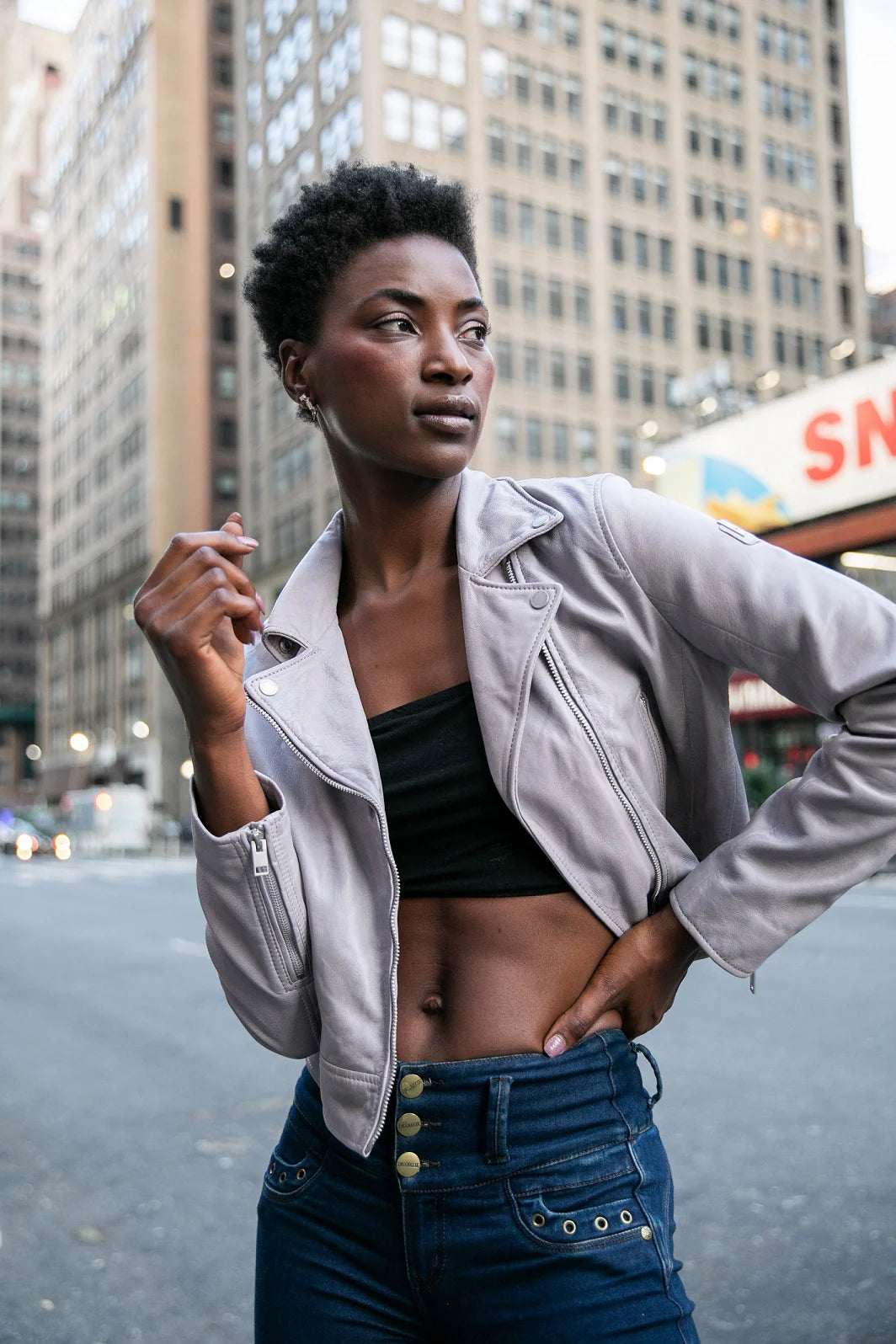 A person with short hair stands confidently on a city street, wearing the chic Julene RF Leather Jacket in Light Lavender from MAURITIUS LEATHER, along with a black crop top and high-waisted jeans that offer a flattering fit. Tall buildings and urban scenery form the blurred background.