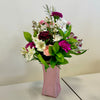 Introducing the "Based in Pink" bouquet by Posies Flowers & Fashion: A bouquet of vibrant flowers arranged in a pink vase. The bouquet includes white daisies, pink carnations, and dark pink carnations, along with assorted greenery. While substitutions may occur based on availability, the same delicate charm is always maintained. The vase sits on a light-colored surface against a plain white background.