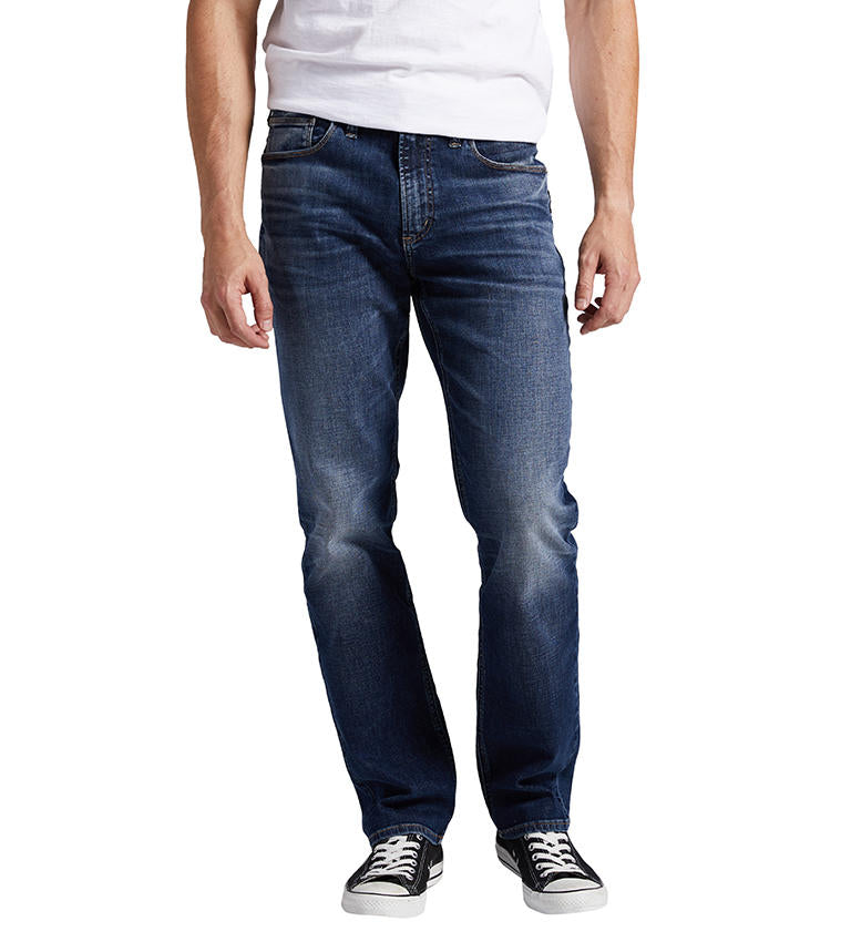 A person stands with arms relaxed at their sides, wearing a white t-shirt, Silver Jeans Co.'s Machray Athletic Fit Straight Leg Jeans in dark blue, and black sneakers with white soles. The person is shown from the shoulder down, without their face visible.