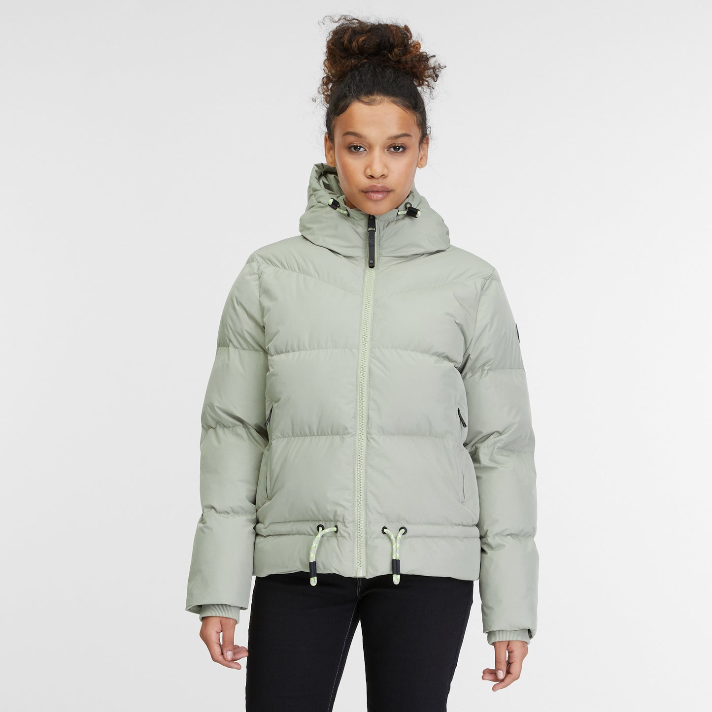 A person with curly hair tied up is wearing a light gray Briony Puffer Jacket 2421-60020 from RAGWEAR, featuring a hood, front zipper, and drawstrings at the waist. Made from vegan materials, it’s both water and windproof. They are also wearing black pants and standing against a plain white background.